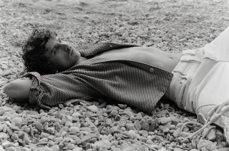 Black and white portrait of a curly-haired male model lying on a rocky beach, wearing an unbuttoned jacket and light pants.