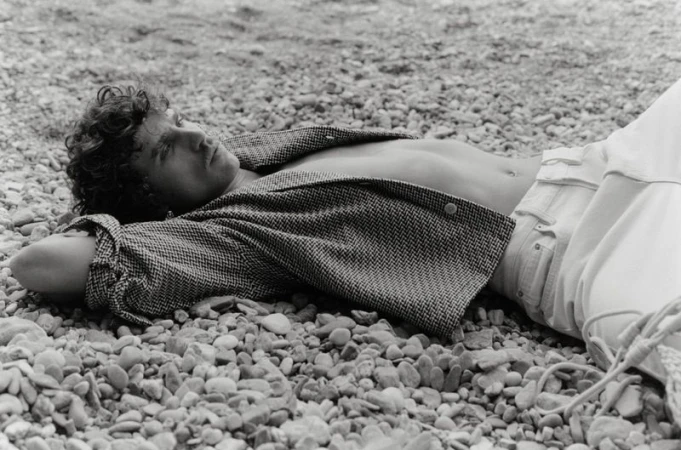 Black and white portrait of a curly-haired male model lying on a rocky beach, wearing an unbuttoned jacket and light pants.
