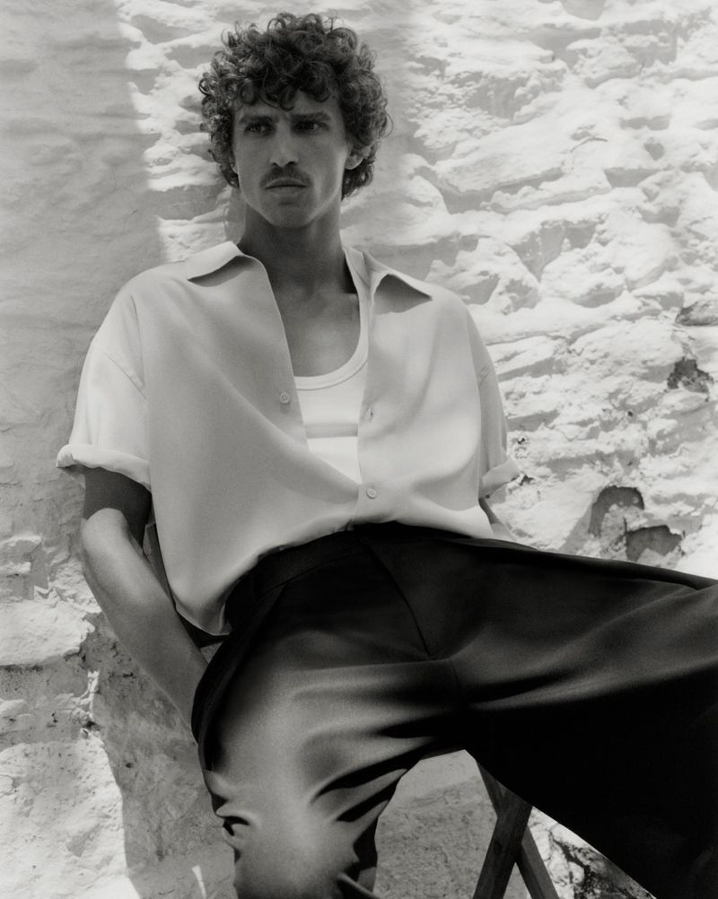 Monochrome portrait of a curly-haired male model posing in an unbuttoned shirt and trousers, leaning against a textured wall.