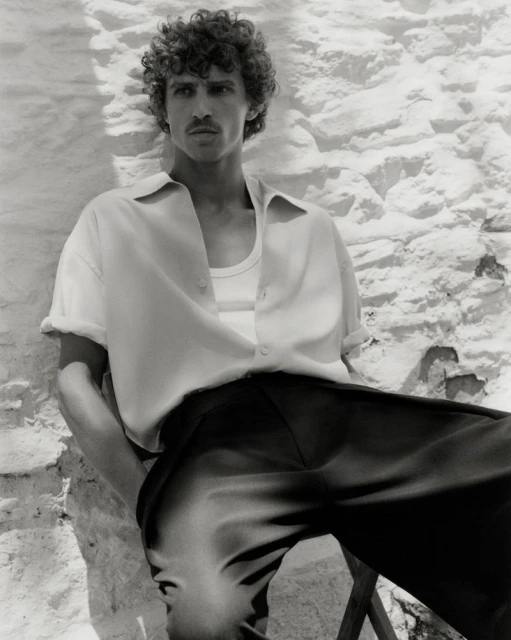 Monochrome portrait of a curly-haired male model posing in an unbuttoned shirt and trousers, leaning against a textured wall.