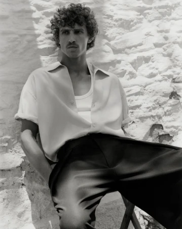 Monochrome portrait of a curly-haired male model posing in an unbuttoned shirt and trousers, leaning against a textured wall.
