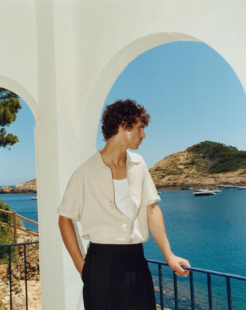 A young adult man in a beige shirt and black pants stands on a white balcony overlooking the blue sea and a green coastal landscape.