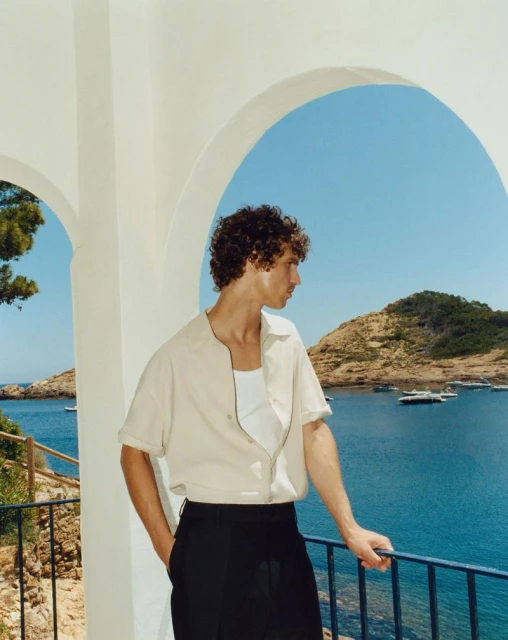 A young adult man in a beige shirt and black pants stands on a white balcony overlooking the blue sea and a green coastal landscape.
