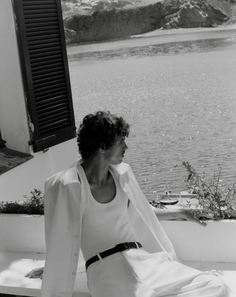 Black and white portrait of a man in a white shirt and pants, sitting and looking out at the water with mountains in background.