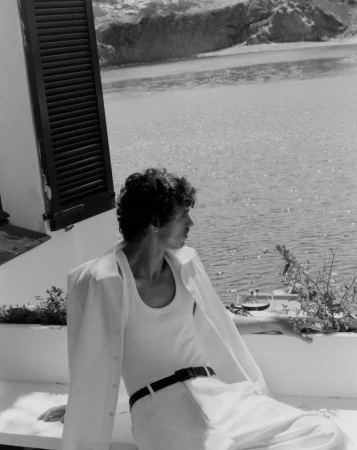 Black and white portrait of a man in a white shirt and pants, sitting and looking out at the water with mountains in background.