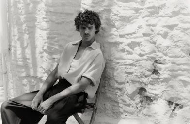 Monochrome portrait of a male model seated on a chair against a textured white stone wall for Zara Sunseeker Edit