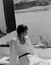 Grzegorz Chanko seated on a sunlit terrace overlooking a coastal bay wearing a white tank top, open shirt, and trousers.