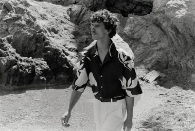 Model Grzegorz Chanko wearing a floral print short-sleeve shirt and light trousers along a rocky coastal shoreline in black and white.