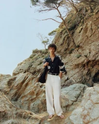 Barefoot man in a black patterned short-sleeve shirt and wide-leg white trousers standing on a rocky cliffside.