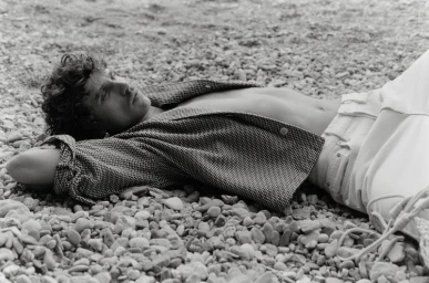 Monochrome fashion photograph of male model Grzegorz Chanko lying on a pebble beach wearing an open patterned shirt.