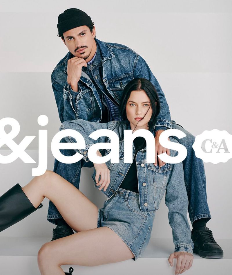 Two fashion models, a man and a woman, are posing together in denim outfits on a white staircase for a clothing brand advertisement.