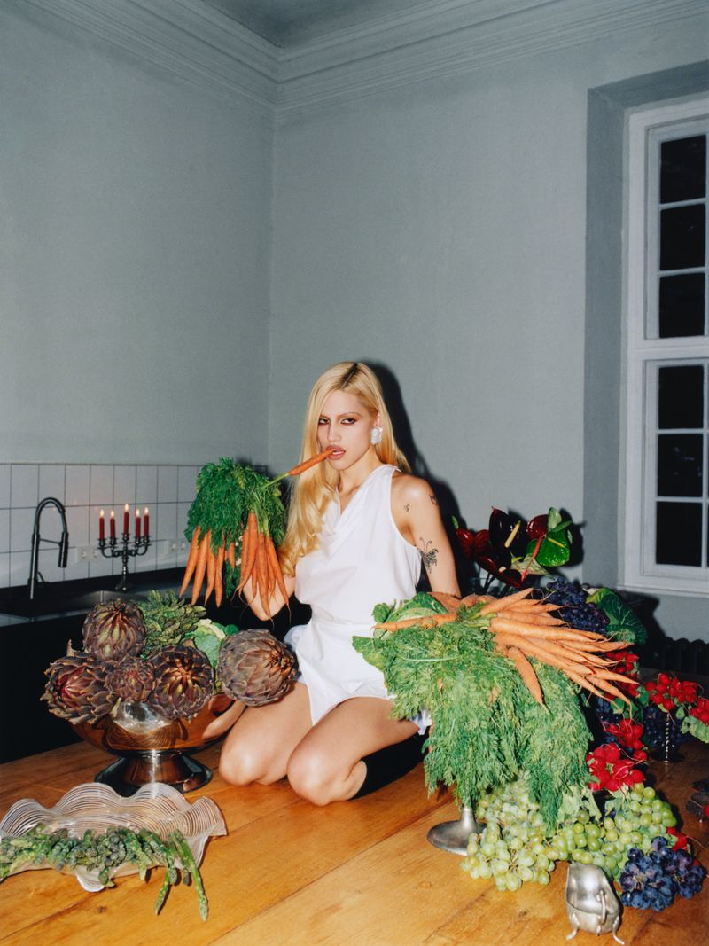 Blonde woman kneeling with carrots and artichokes on a wooden table indoors, lit by natural window light and candlelight.