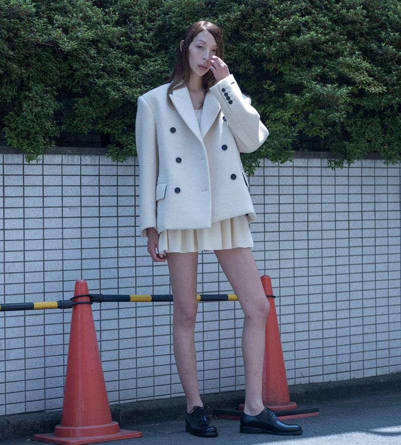 An adult female stands outdoors in a double-breasted cream blazer with a pleated cream skirt and black shoes in an urban setting.