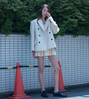 Model Issa Lish wearing a cream oversized double-breasted blazer and matching pleated mini skirt outdoors by a tiled wall.