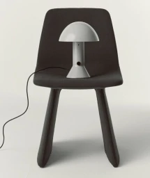 A white dome-shaped table lamp resting on the seat of a modern dark grey upholstered chair against a studio background.