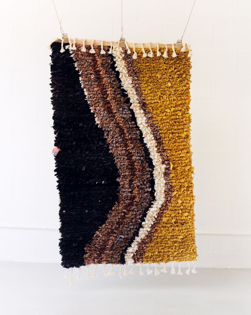Abstract wall art featuring a woven textile hanging with varied colors: black, brown, tan, and cream, suspended against white wall.