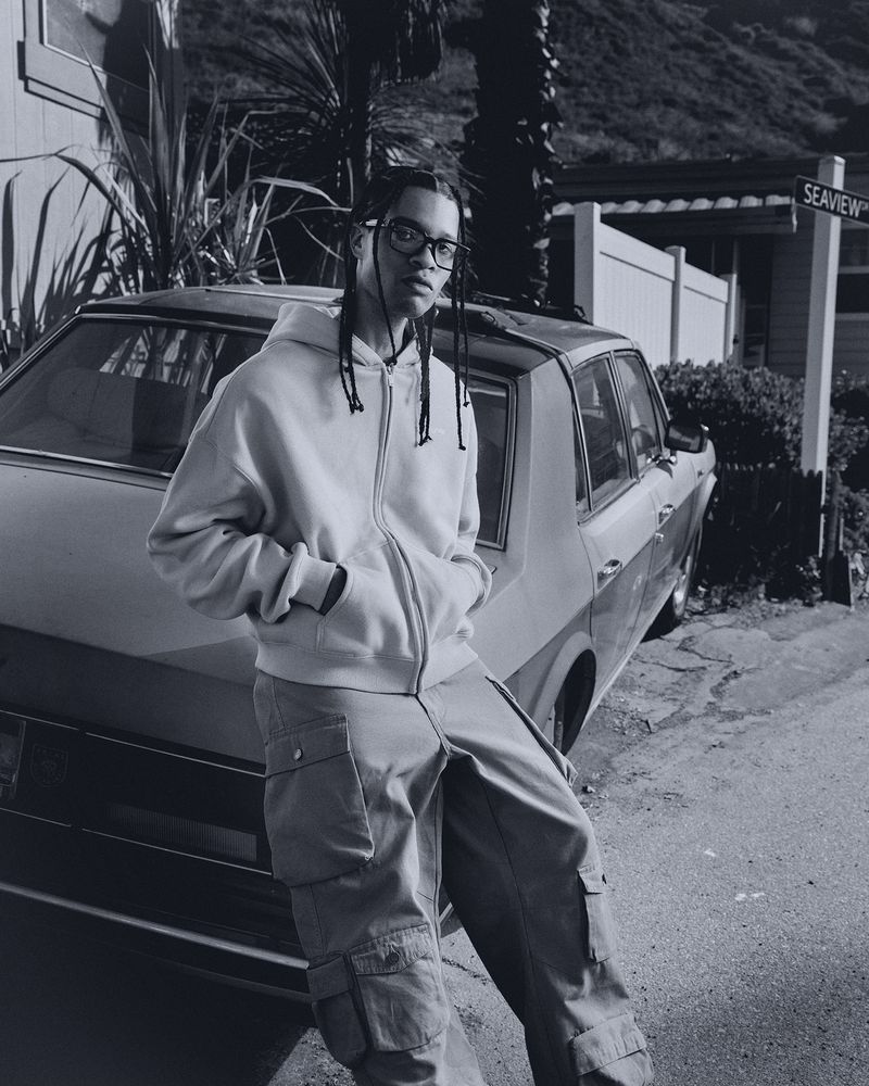 A black and white medium shot of a man with long braids leaning against a car in front of a house on a tree-lined street.
