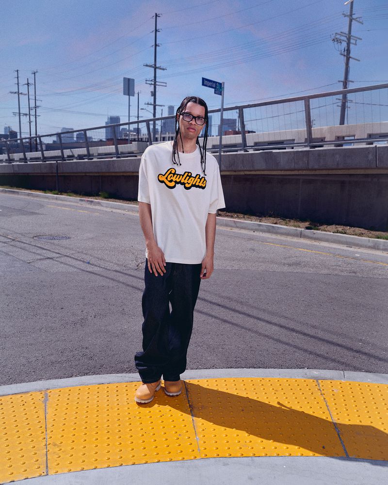 Man in white t-shirt with 'Lowlight' logo poses on street with buildings and utility poles against a blue sky backdrop on a sunny day.