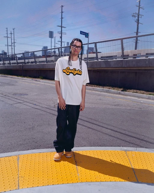 Man in white t-shirt with 'Lowlight' logo poses on street with buildings and utility poles against a blue sky backdrop on a sunny day.