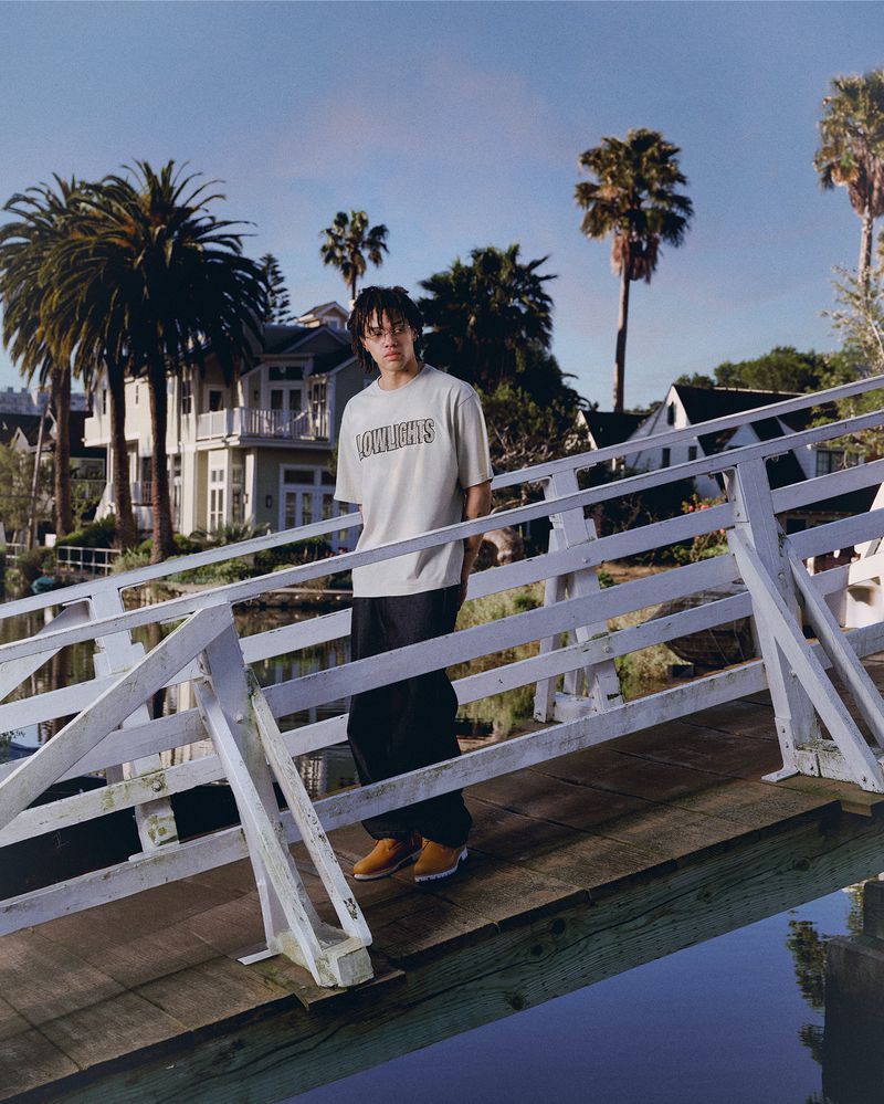 An adult stands on a pier wearing a t-shirt and wide-legged pants, posing outdoors with water, palm trees, and houses in the background.