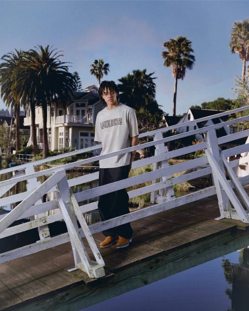 An adult stands on a pier wearing a t-shirt and wide-legged pants, posing outdoors with water, palm trees, and houses in the background.