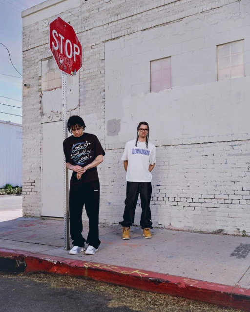 Two people stand near a stop sign in an urban setting, wearing casual clothes and sneakers, under natural lighting.