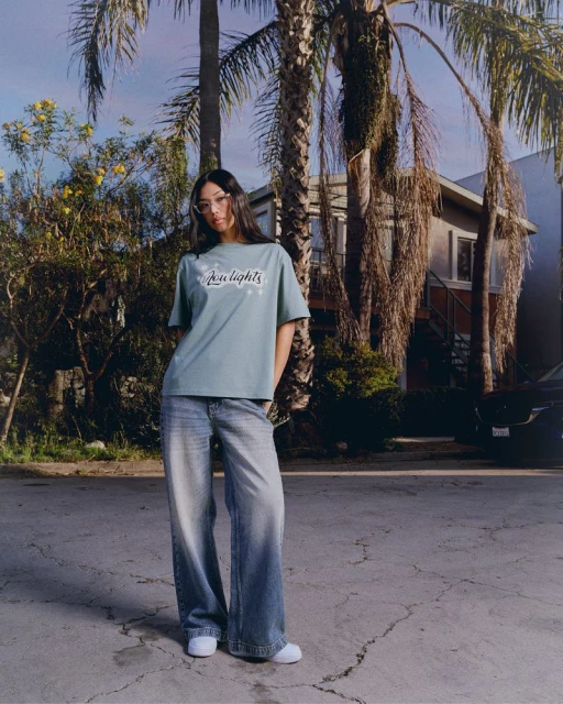 An adult woman stands on a cracked street in casual clothing, including a graphic tee and wide-leg jeans, with palm trees and a house behind.
