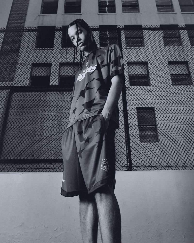Monochrome portrait of a young adult in sportswear posing outdoors against a wire fence and apartment building backdrop.