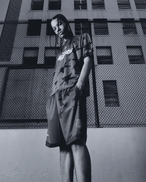 Monochrome portrait of a young adult in sportswear posing outdoors against a wire fence and apartment building backdrop.