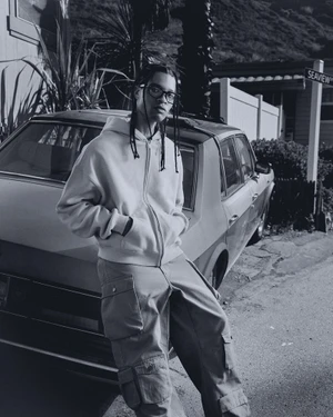 Young man with braided hair and glasses leaning against a parked car wearing a zip hoodie and cargo pants.