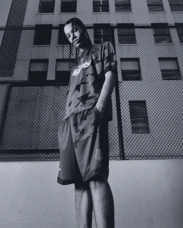 Julian Trowe posing in a graphic star-print jersey and shorts set in front of a chain-link fence and building.