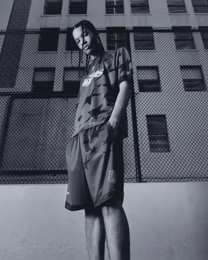 Julian Trowe posing in a graphic star-print jersey and shorts set in front of a chain-link fence and building.