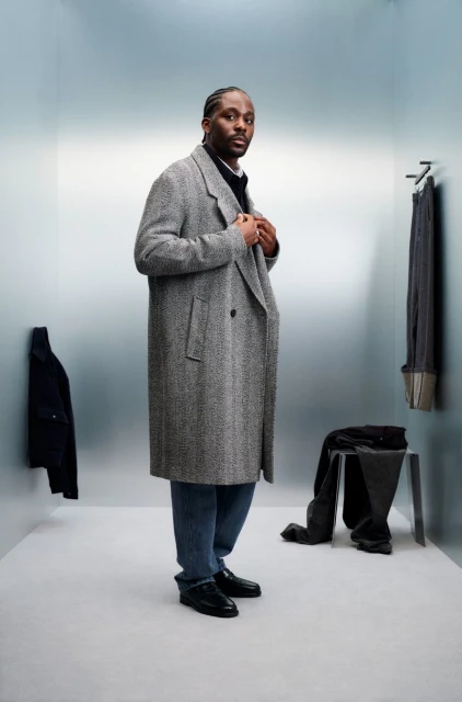 Man in grey herringbone overcoat, jeans, and black loafers stands in a photo studio with blue wall, and clothing on display.