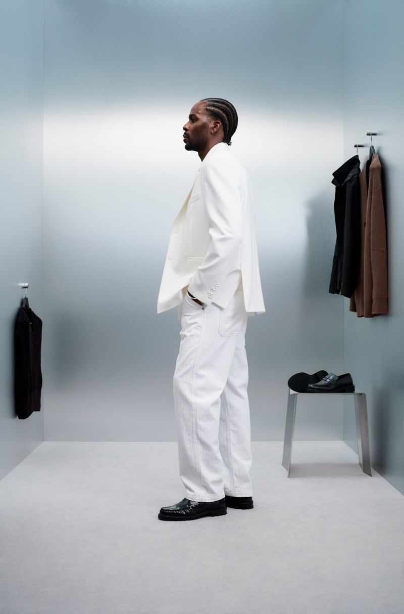 Side profile of a black male adult in a white suit in a photo studio with a metal backdrop and black loafers on, minimal aesthetic.