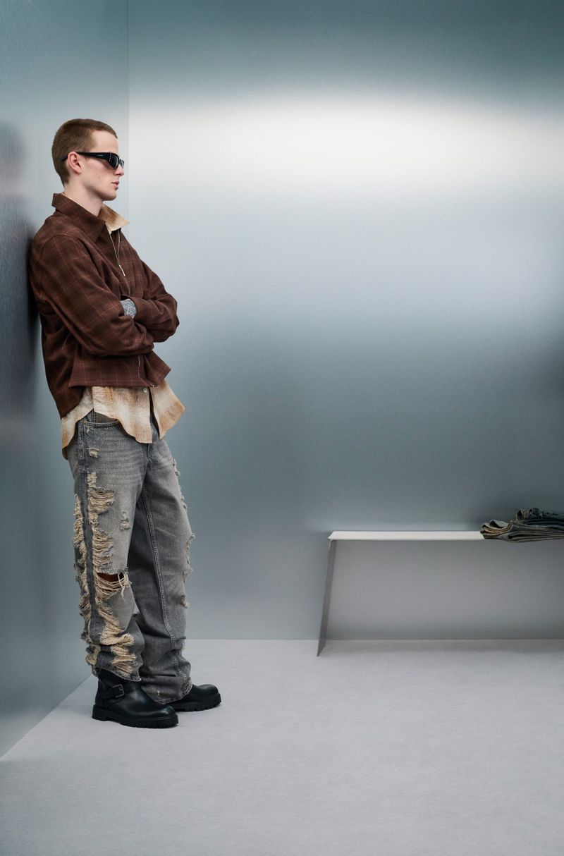 A male model with sunglasses stands against a wall in a brown jacket and distressed jeans in a fashion editorial shot indoors.