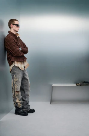 A male model with sunglasses stands against a wall in a brown jacket and distressed jeans in a fashion editorial shot indoors.