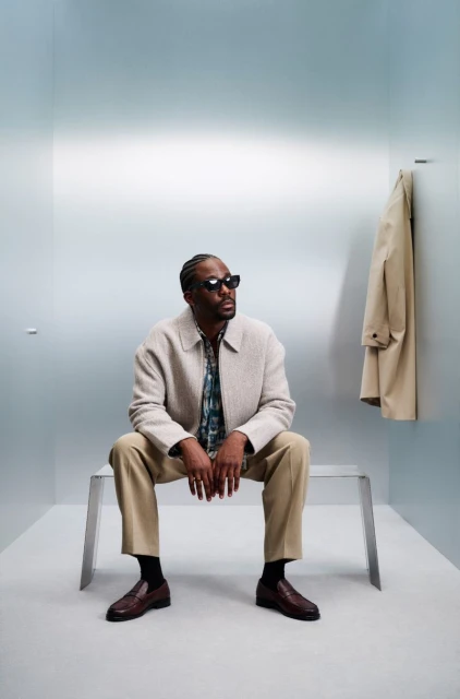 A stylish Black man is seated on a metal bench in a pale blue room, wearing a beige jacket, tan pants, and sunglasses, with a tan coat hanging.