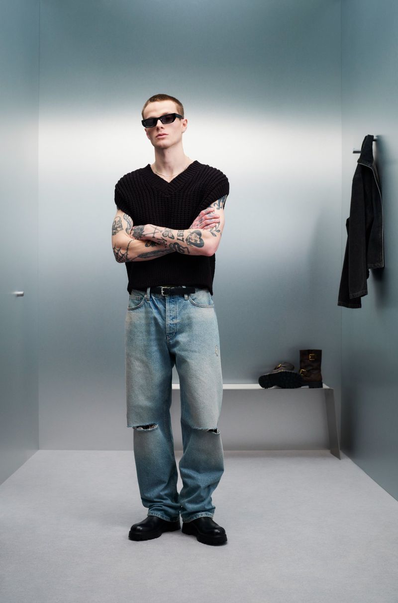 Man in black vest, ripped jeans, and sunglasses stands in a minimalist room. Fashion editorial with industrial backdrop and neutral tones.