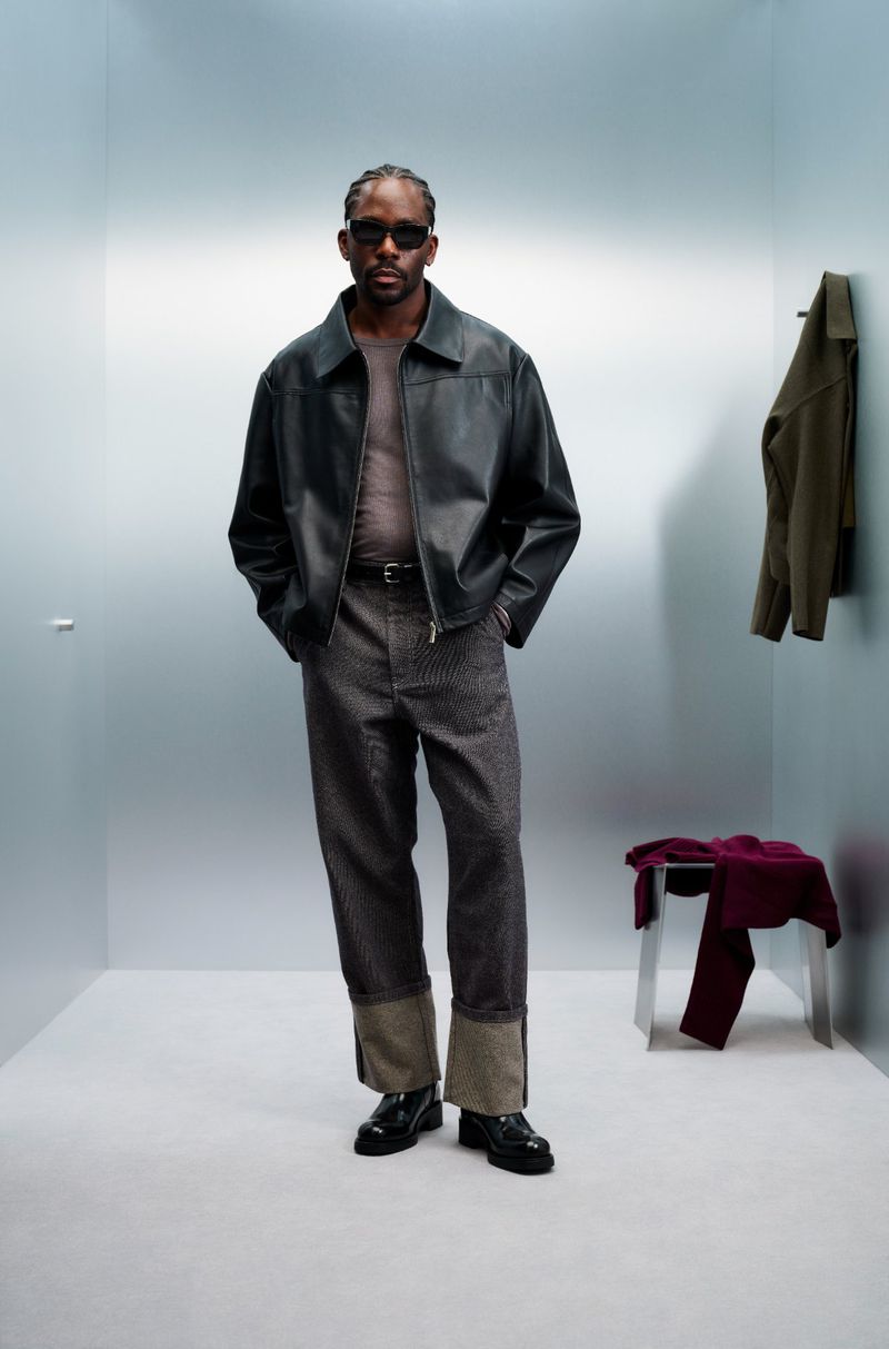 A stylish dark-skinned man poses indoors wearing a black leather jacket, grey textured pants, and black sunglasses, exuding confidence.