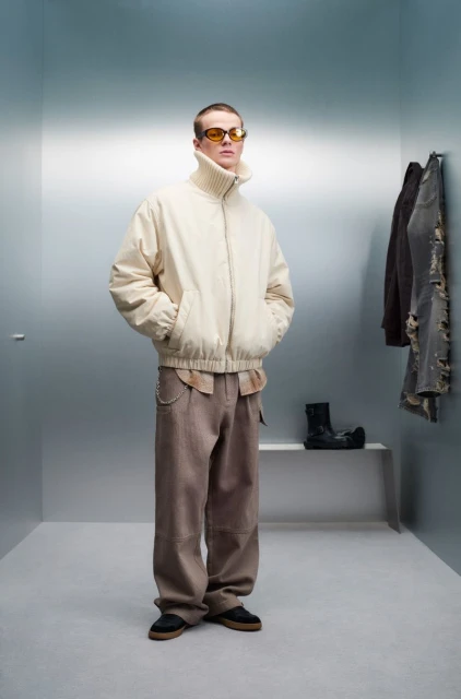 Full-length studio shot of a man in a beige jacket, brown pants, and yellow sunglasses standing in a corner with hanging clothes.
