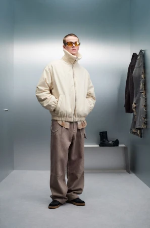 Full-length studio shot of a man in a beige jacket, brown pants, and yellow sunglasses standing in a corner with hanging clothes.