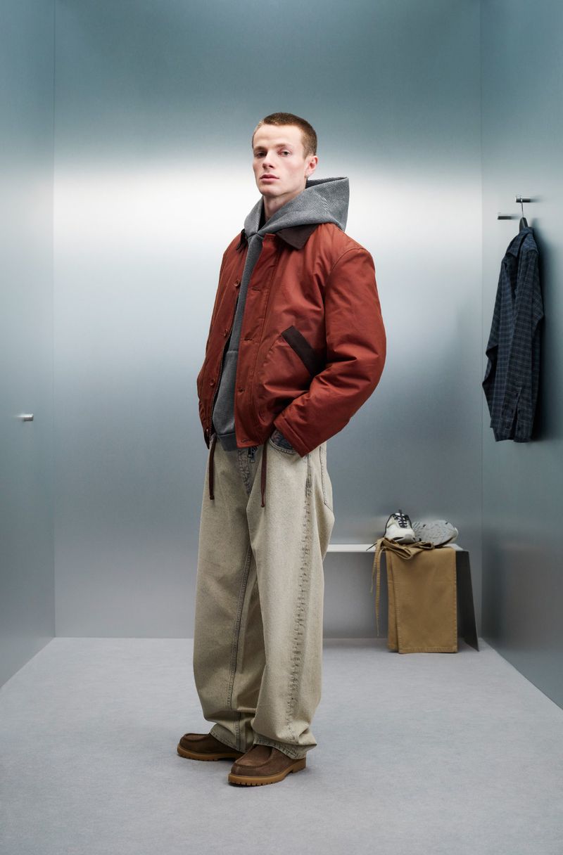 Full-length shot of a young adult male wearing a rust-colored bomber jacket, grey hoodie, beige jeans, and brown shoes in a studio setting.