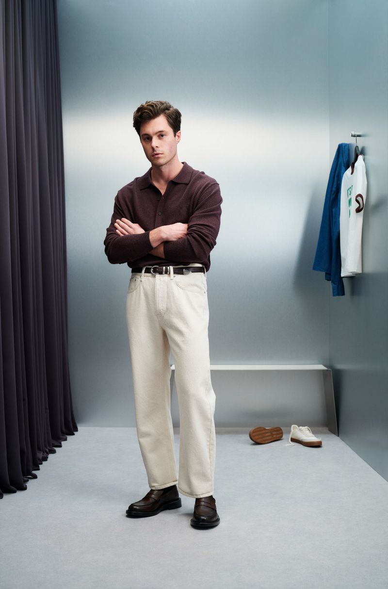 Man in a brown collared shirt and cream pants standing indoors with his arms crossed. Clothes hanging on the wall in the background.