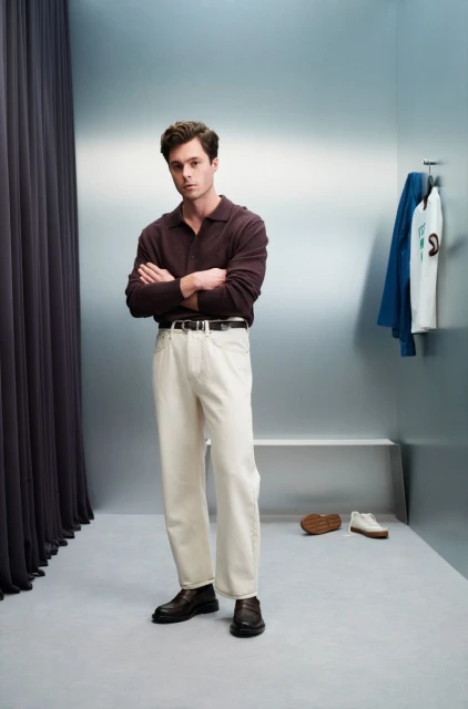 Man in a brown collared shirt and cream pants standing indoors with his arms crossed. Clothes hanging on the wall in the background.