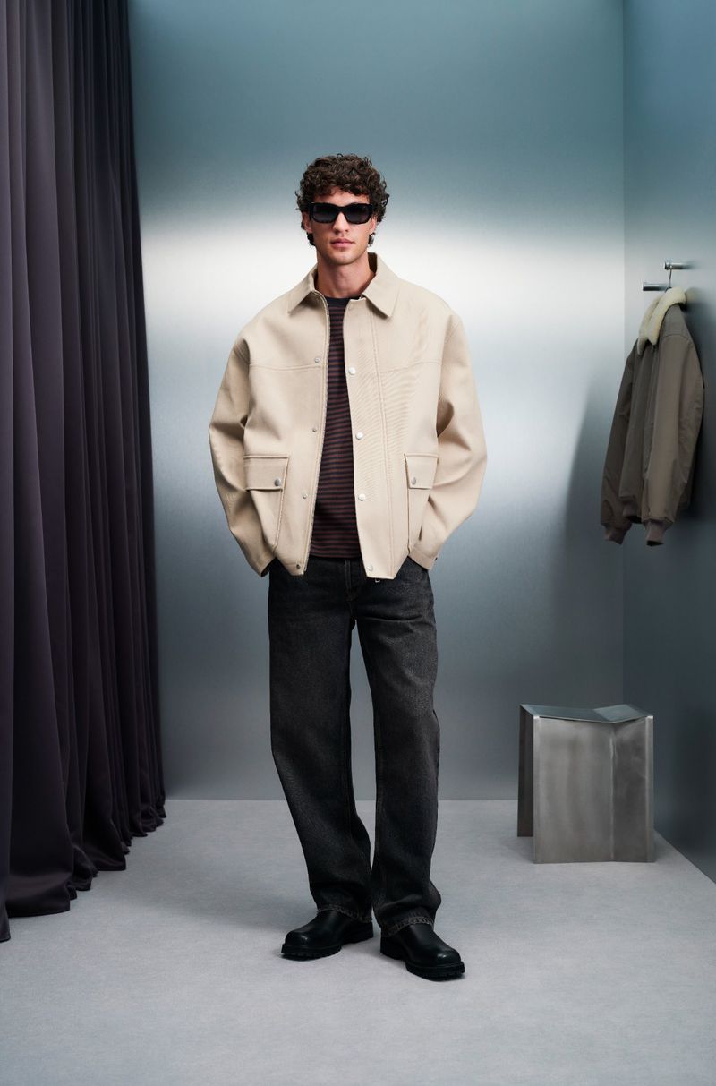 Full-shot of a trendy man wearing a beige jacket and dark jeans in a photo studio with modern lighting and a minimalist metal side table.