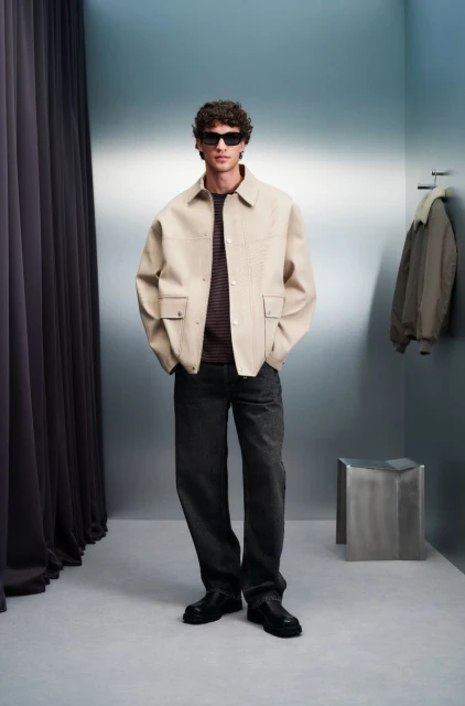 Full-shot of a trendy man wearing a beige jacket and dark jeans in a photo studio with modern lighting and a minimalist metal side table.