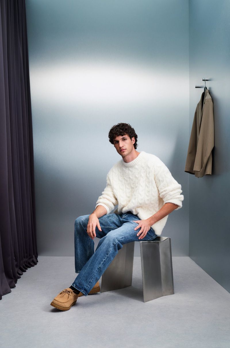 Adult male seated on a metal stool. He is wearing a white sweater, blue jeans, and brown shoes, photographed indoors against a gradient wall.