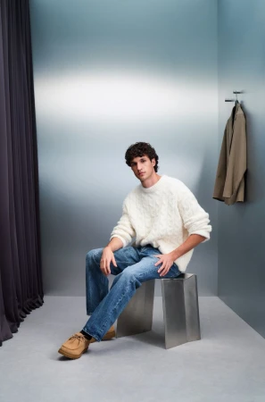 Adult male seated on a metal stool. He is wearing a white sweater, blue jeans, and brown shoes, photographed indoors against a gradient wall.