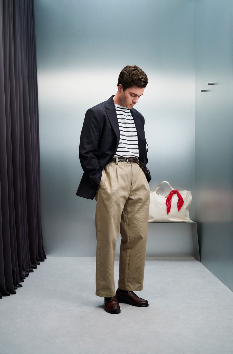 Man in a blazer, striped shirt, and khaki pants stands in a minimalist studio; he is looking down with his hands in his pockets.