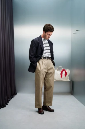Man in a blazer, striped shirt, and khaki pants stands in a minimalist studio; he is looking down with his hands in his pockets.